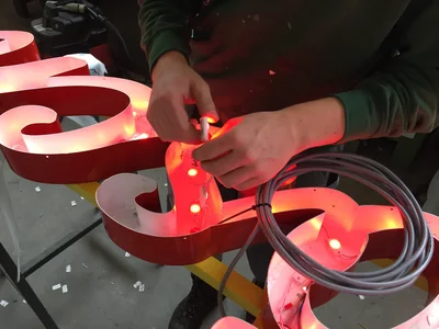 A worker fits LED modules inside a red channel letter on the bench.